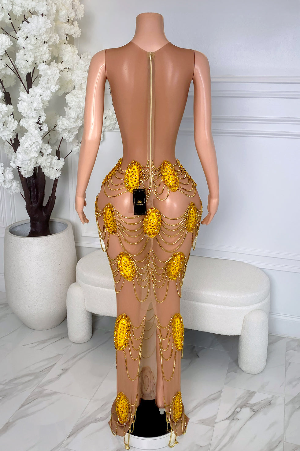 Back view of nude illusion mesh maxi dress with gold rhinestone appliqué, draped gold chain accents, open back, and zipper closure