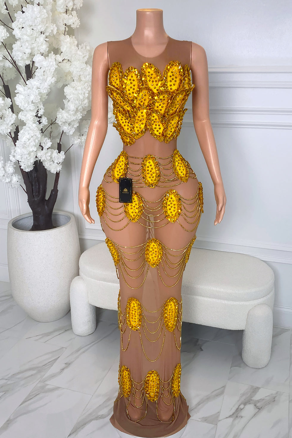 Front view of nude illusion mesh maxi dress with gold rhinestone appliqué, draped gold chain accents, sleeveless bodice, and body-hugging fit