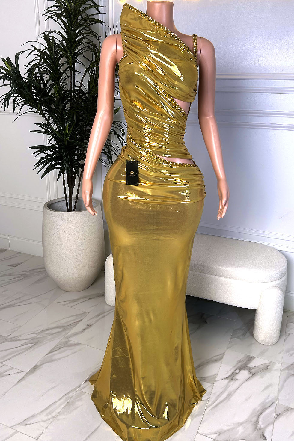 Front view of a metallic gold one shoulder evening gown with ruched detailing, side cutout, embellished trim, and floor-length silhouette