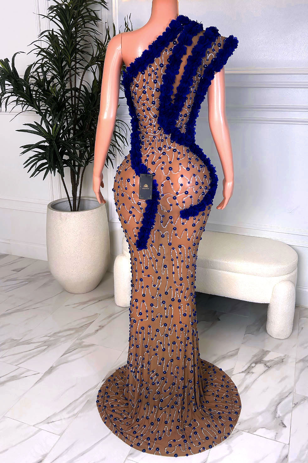 Nude illusion mesh mermaid maxi gown covered in royal blue rhinestones with bold 3D blue floral trim and a sweeping train, back view.